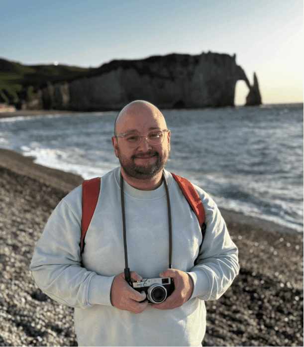 Aleksei on a pebble beach below a chalk headland, a camera around his neck.